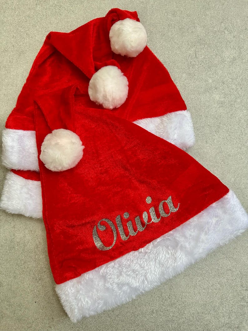 May include: Three red velvet Santa hats with white fur trim and white pom-poms. The bottom hat has the name "Olivia" embroidered in silver glitter.