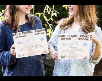 May include: Two women hold up certificates that say "Baby Dundie Award" with one saying "Nomination" and the other saying "Dundie Winner". The certificates are printed on a cream background with brown text and blue accents.