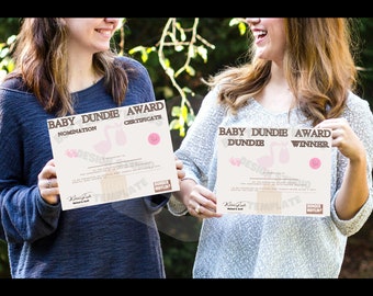 May include: Two women hold up pink and white baby shower certificates that say "Baby Dundie Award" with one certificate saying "Nomination" and the other saying "Dundie Winner".