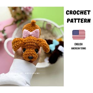 Könnte beinhalten: Ein braunes, gehäkeltes Plüschhund-Spielzeug mit einer hellrosa Schleife auf dem Kopf, schwarzen Kunststoffaugen und einer schwarzen Nase. Der Text CROCHET PATTERN, eine US-Flaggen-Ikone und ENGLISH AMERICAN TERMS sind rechts abgebildet.