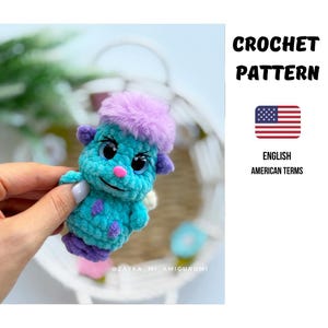 Könnte beinhalten: Eine kleine, türkisfarbene Häkelfigur mit einem flauschigen lila Schopf auf dem Kopf, lila Füßen und lila Flecken. Sie hat große, dunkle, ausdrucksstarke Augen und eine kleine rosa Nase. Der Text CROCHET PATTERN und eine US-Flagge sind zu sehen.
