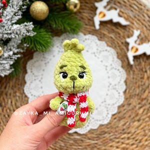 May include: A green crocheted Grinch doll with black eyes and a red and white striped scarf. The doll is holding a small tag. The background includes Christmas decorations and the text "@ZAYKA_MI_AMIGURUMI".