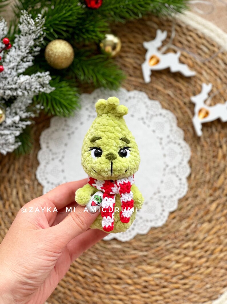 May include: A green crocheted Grinch doll with black eyes and a red and white striped scarf. The doll is holding a small tag. The background includes Christmas decorations and the text "@ZAYKA_MI_AMIGURUMI".