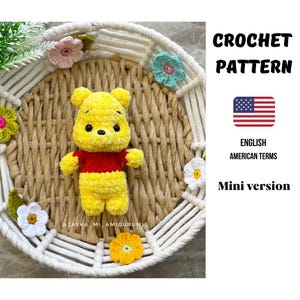 May include: A crocheted yellow bear with a red shirt, surrounded by crocheted flowers in a woven basket. The text "CROCHET PATTERN" and "ENGLISH AMERICAN TERMS" are visible. The bear is a mini version.