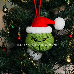 May include: A green crocheted Christmas ornament in the shape of a character's face, wearing a red Santa hat with white trim and a white pom-pom. The ornament hangs on a Christmas tree. The text "@ZAYKA_MI_AMIGURUMI" is visible.
