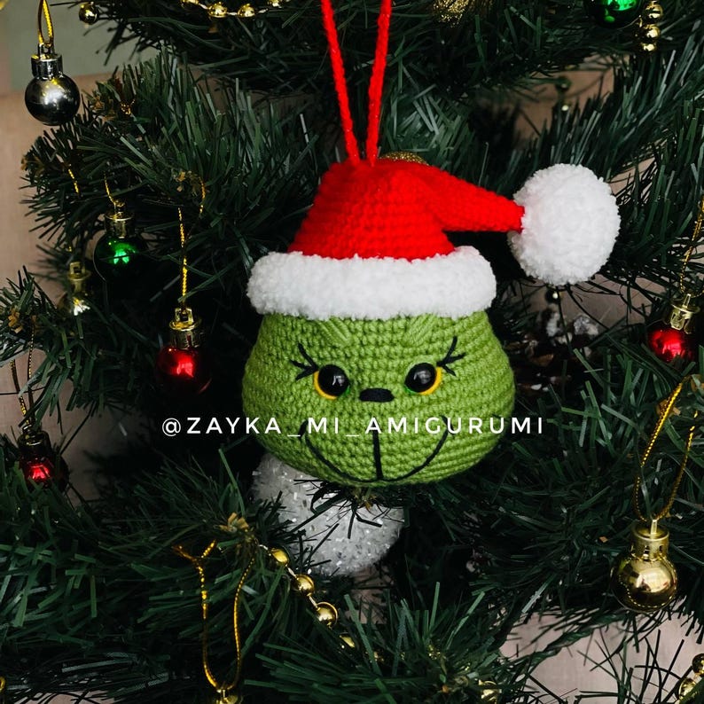 May include: A green crocheted Christmas ornament in the shape of a character's face, wearing a red Santa hat with white trim and a white pom-pom. The ornament hangs on a Christmas tree. The text "@ZAYKA_MI_AMIGURUMI" is visible.