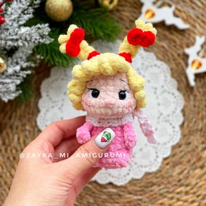 May include: A handmade crochet doll with a pink dress, yellow hair, and red accents. The doll has a white scarf and is holding a nail with a green graphic. The doll is a Christmas-themed amigurumi.