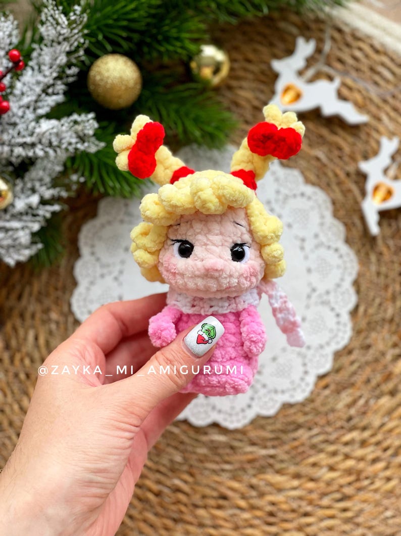 May include: A handmade crochet doll with a pink dress, yellow hair, and red accents. The doll has a white scarf and is holding a nail with a green graphic. The doll is a Christmas-themed amigurumi.