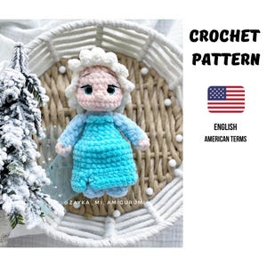 Könnte beinhalten: Gehäkelte Puppe in hellblauem Kleid und weißen Haaren, platziert auf einem geflochtenen Korb. Der Text lautet "CROCHET PATTERN" und "ENGLISH AMERICAN TERMS", mit einer amerikanischen Flagge.