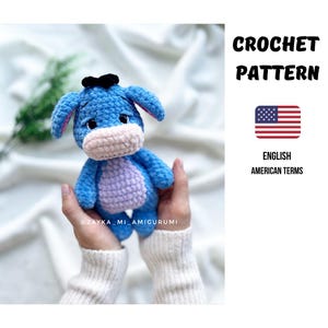 Könnte beinhalten: Ein blaues Häkel-Esel-Amigurumi mit schwarzer Schleife, rosa Ohren und hellrosa Schnauze. Der Esel hat einen lila Bauch. Das Bild enthält den Text "CROCHET PATTERN" und "ENGLISH AMERICAN TERMS".
