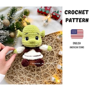 Könnte beinhalten: Gehäkelte Shrek-Puppe mit grünem Kopf, weißem Hemd und brauner Weste. Die Puppe hat schwarze Augen und hält einen Weihnachtsschmuck. Das Bild enthält den Text "CROCHET PATTERN" und "ENGLISH AMERICAN TERMS".