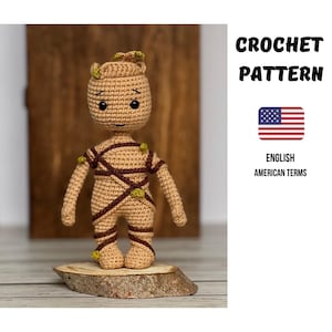 Puede incluir: Figura de ganchillo beige con detalles marrones, que representa un personaje. Tiene una cabeza redonda, extremidades pequeñas y se muestra sobre una base de madera. El texto "CROCHET PATTERN" y "ENGLISH AMERICAN TERMS" también es visible.