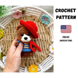 Könnte beinhalten: Ein gehäkelter brauner Teddybär mit rotem Hut, blauem Pullover mit gelben Streifen und rotem Schal. Der Bär hat eine weiße Schnauze und schwarze Augen. Das Bild enthält den Text "CROCHET PATTERN" und "ENGLISH AMERICAN TERMS".