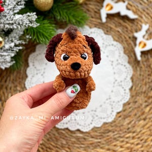 May include: A handmade brown amigurumi dog with dark brown ears and a mohawk. The dog has white eyes and a black nose. The amigurumi is held in a hand, with a white doily and Christmas decorations in the background. The text "@ZAYKA_MI_AMIGURUMI" is visible.