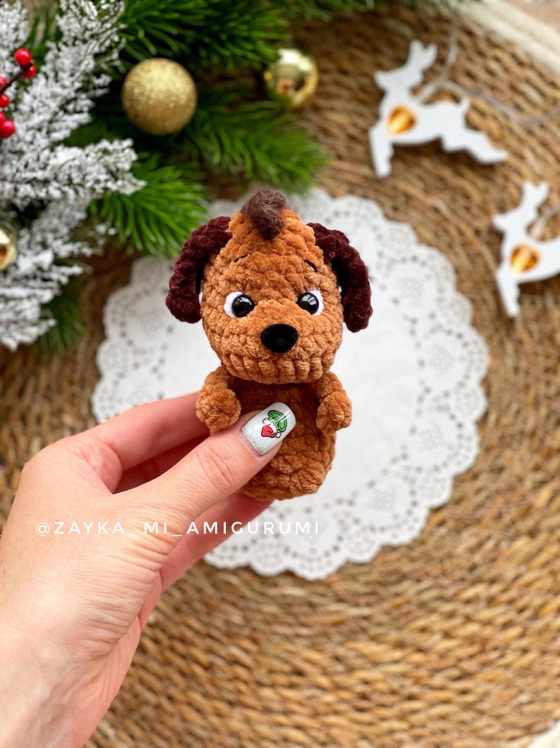 May include: A handmade brown amigurumi dog with dark brown ears and a mohawk. The dog has white eyes and a black nose. The amigurumi is held in a hand, with a white doily and Christmas decorations in the background. The text "@ZAYKA_MI_AMIGURUMI" is visible.