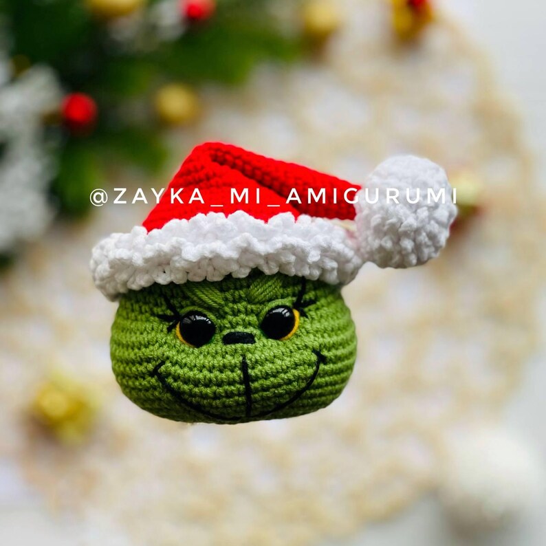 May include: A handmade crocheted Grinch head, complete with a red and white Santa hat. The Grinch's green face features black eyes and a stitched smile. The image includes the text "@ZAYKA_MI_AMIGURUMI".