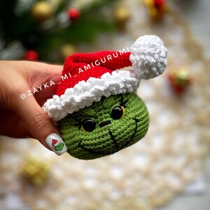 May include: A crocheted green character wearing a red Santa hat with white trim and a pom-pom. The character has black eyes and a stitched smile. The text "ZAYKA_MI_AMIGURUMI" is visible.