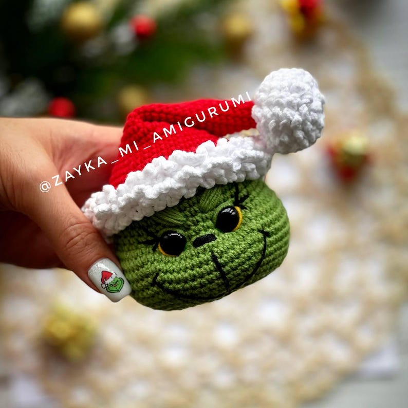 May include: A crocheted green character wearing a red Santa hat with white trim and a pom-pom. The character has black eyes and a stitched smile. The text "ZAYKA_MI_AMIGURUMI" is visible.