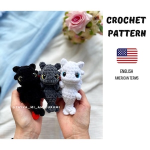 Könnte beinhalten: Drei gehäkelte Drachenspielzeuge in Schwarz, Grau und Weiß, jeweils mit großen Augen und kleinen Flügeln. Der Text "CROCHET PATTERN" und eine amerikanische Flagge sind sichtbar. Der Text "ENGLISH AMERICAN TERMS" ist ebenfalls sichtbar.