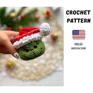May include: A green crocheted Grinch figure wearing a red and white Santa hat. The amigurumi has a smiling face with black eyes and a stitched mouth. The image includes text that says "CROCHET PATTERN" and "ENGLISH AMERICAN TERMS".