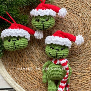 May include: Handmade crochet Christmas decorations. The green character wears a red and white Santa hat and a striped scarf. The ornaments are displayed on a woven surface. The ornaments have a smiling face.