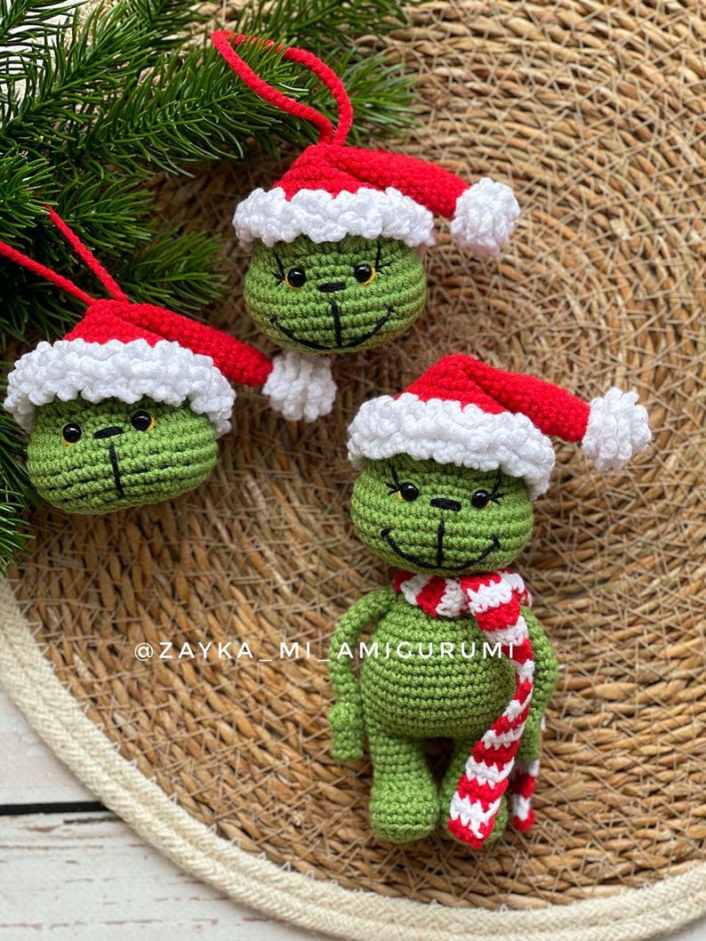 May include: Handmade crochet Christmas decorations. The green character wears a red and white Santa hat and a striped scarf. The ornaments are displayed on a woven surface. The ornaments have a smiling face.