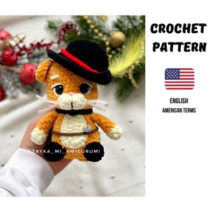 Puede incluir: Amigurumi de gato naranja tejido a ganchillo con un sombrero negro con una banda roja. El gato tiene bigotes blancos, una barriga color crema y un cinturón marrón. El texto "CROCHET PATTERN" y "ENGLISH AMERICAN TERMS" son visibles.