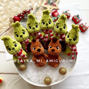 May include: Crocheted green figures resembling a character with red and white striped scarves, alongside brown crocheted dog figures. The figures are arranged in a circular pattern, with gold glitter balls. The text "@ZAYKA_MI_AMIGURUMI" is visible.