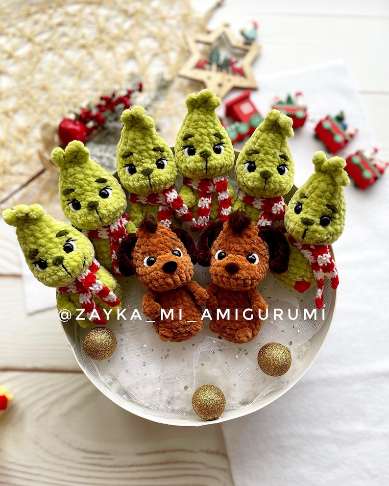 May include: Crocheted green figures resembling a character with red and white striped scarves, alongside brown crocheted dog figures. The figures are arranged in a circular pattern, with gold glitter balls. The text "@ZAYKA_MI_AMIGURUMI" is visible.
