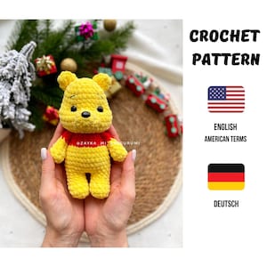 Könnte beinhalten: Eine gelbe gehäkelte Winnie-the-Pooh-Puppe mit roten Akzenten um den Hals. Die Puppe wird in zwei Händen gehalten. Das Bild enthält auch den Text "CROCHET PATTERN", "ENGLISH AMERICAN TERMS" und "DEUTSCH".