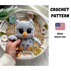 Könnte beinhalten: Ein gehäkelter Eulen-Amigurumi in Grautönen, mit großen, ausdrucksstarken Augen und einem orangefarbenen Schnabel. Die Eule wird in einer Hand gehalten, mit dem Text "CROCHET PATTERN" und "ENGLISH AMERICAN TERMS" sichtbar. Die Eule ist von dekorativen Blumen umgeben.