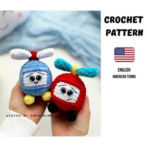 Könnte beinhalten: Zwei gehäkelte Helikopter-Spielzeuge, einer blau und einer rot, jeweils mit weißen und blauen Propellern und einem lächelnden Gesicht. Das Bild enthält den Text "CROCHET PATTERN" und "ENGLISH AMERICAN TERMS" mit einer amerikanischen Flagge.