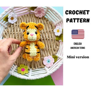 Könnte beinhalten: Ein gehäkeltes Tiger-Spielzeug mit orangefarbenen und schwarzen Streifen, einer rosa Nase und schwarzen Augen. Das Spielzeug wird in einem geflochtenen Korb mit gehäkelten Blumen präsentiert. Das Bild enthält den Text "CROCHET PATTERN" und "Mini version".