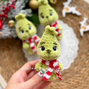 May include: Handmade green amigurumi doll with a red and white scarf. The doll has black eyes and a stitched mouth. The doll is a Christmas-themed character. The text "@ZAYKA_ML_AMIGURUMI" is visible.