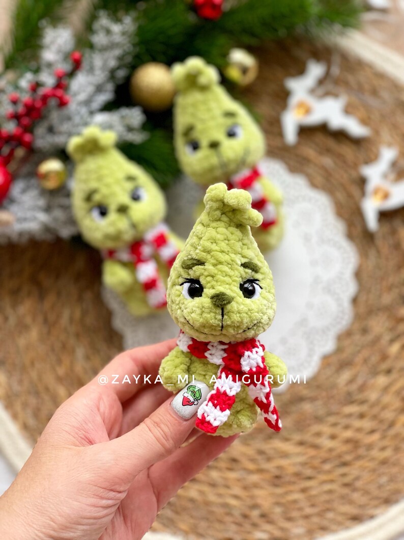 May include: Handmade green amigurumi doll with a red and white scarf. The doll has black eyes and a stitched mouth. The doll is a Christmas-themed character. The text "@ZAYKA_ML_AMIGURUMI" is visible.