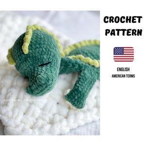 Könnte beinhalten: Ein grünes gehäkeltes Drachenspielzeug mit gelben Akzenten, das auf einer weißen, flauschigen Oberfläche liegt. Das Bild enthält den Text "CROCHET PATTERN" und "ENGLISH AMERICAN TERMS" mit einer US-Flagge. Der Drache ist ein handgemachtes Amigurumi.