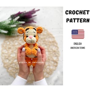 Könnte beinhalten: Ein gehäkeltes orange-cremefarbenes Tiger-Amigurumi mit schwarzen Streifen, einer rosa Nase und großen Ohren. Der Text "CROCHET PATTERN" wird in Schwarz angezeigt, zusammen mit der amerikanischen Flagge und den Worten "ENGLISH AMERICAN TERMS".
