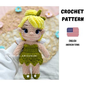 Könnte beinhalten: Eine handgefertigte Häkelpuppe mit einem grünen Kleid, gelben Haaren und weißen Flügeln. Die Puppe hat blaue Augen und lange Wimpern. Der Text "CROCHET PATTERN" wird mit einer amerikanischen Flagge und den Worten "ENGLISH AMERICAN TERMS" angezeigt.
