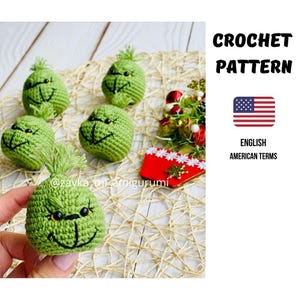Puede incluir: Varios adornos verdes tejidos a ganchillo, cada uno con una cara sonriente, detalles negros y ojos amarillos. Están dispuestos sobre una superficie tejida. El texto indica "CROCHET PATTERN" y "ENGLISH AMERICAN TERMS".