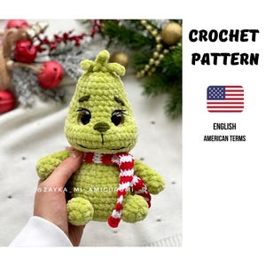 May include: A handmade green crochet Grinch doll wearing a red and white striped scarf. The doll has large black eyes and a stitched mouth. The image includes text that says "CROCHET PATTERN" and "ENGLISH AMERICAN TERMS".