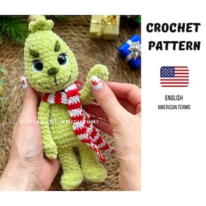 Puede incluir: Un personaje verde tejido a ganchillo con una bufanda a rayas rojas y blancas. El personaje tiene una cara sonriente con ojos negros y una nariz pequeña. La imagen incluye el texto "CROCHET PATTERN" y "ENGLISH AMERICAN TERMS" con una bandera estadounidense.