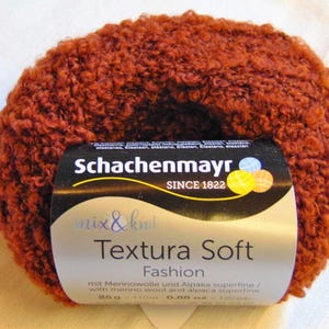 Könnte beinhalten: Ein Knäuel rostfarbener Wolle mit einer strukturierten, geschlungenen Optik. Die Wolle ist mit "Schachenmayr Textura Soft Fashion" mit dem Text "SINCE 1822" gekennzeichnet. Das Etikett weist auch darauf hin, dass die Wolle aus Merinowolle und Alpaka superfine besteht.