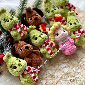 May include: A collection of handmade crochet dolls, including green characters with red and white striped scarves and brown dog dolls. The dolls are arranged on a textured surface with greenery, perfect for holiday decor. The text "ZAYKA_MI_AMIGURUMI" is visible.