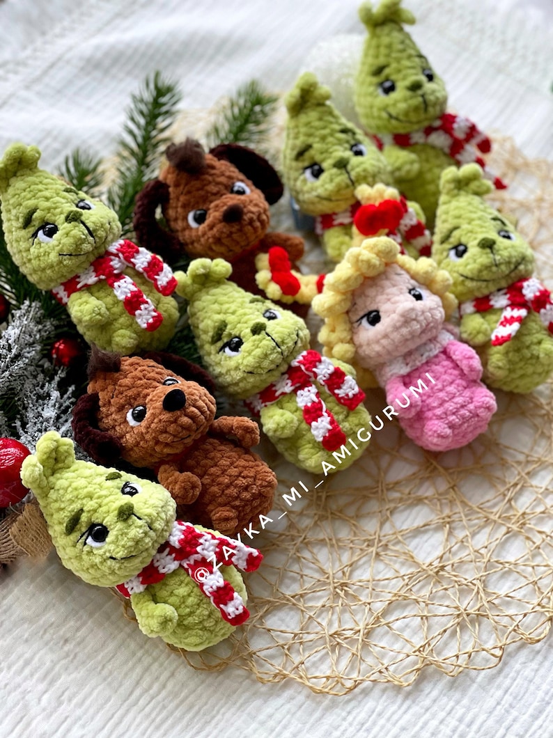 May include: A collection of handmade crochet dolls, including green characters with red and white striped scarves and brown dog dolls. The dolls are arranged on a textured surface with greenery, perfect for holiday decor. The text "ZAYKA_MI_AMIGURUMI" is visible.