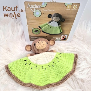 Op de afbeelding: Een gehaakte Kiwi Monkey Lovey, met een bruine apenkop en een groene en bruine kiwischijfdeken. Het product wordt gepresenteerd op een witte, pluizige ondergrond, met een doos op de achtergrond met de tekst "Anchor Baby Pure Cotton".