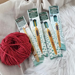 Könnte beinhalten: Ein Knäuel dunkelrotes Garn neben vier Clover Soft Touch Häkelnadeln in ihrer Originalverpackung. Die Häkelnadeln haben goldfarbene Griffe mit dunklen ovalen Einsätzen und sind mit den Größen 5,0 mm, 4,5 mm, 2,25 mm und 0,50 mm beschriftet.