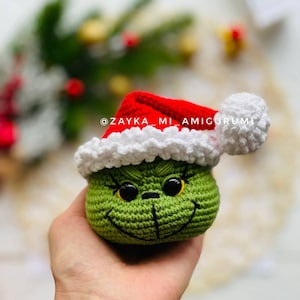 May include: A crocheted green Grinch head wearing a red Santa hat with white trim and a white pom-pom. The Grinch has black eyes, a stitched smile, and eyelashes. The text "@ZAYKA_MI_AMIGURUMI" is visible.