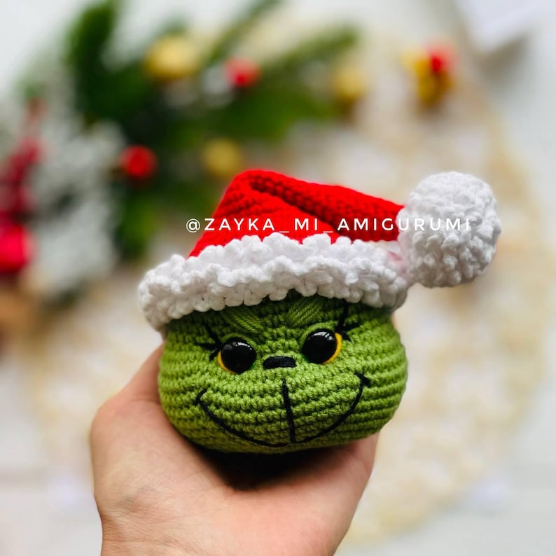 May include: A crocheted green Grinch head wearing a red Santa hat with white trim and a white pom-pom. The Grinch has black eyes, a stitched smile, and eyelashes. The text "@ZAYKA_MI_AMIGURUMI" is visible.