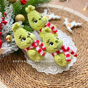 May include: Three handmade green crochet figures with black eyes and red and white striped scarves. The figures are arranged on a white lace doily, with Christmas ornaments and greenery in the background. The text "@ZAYKA_MI_AMIGURUMI" is visible.