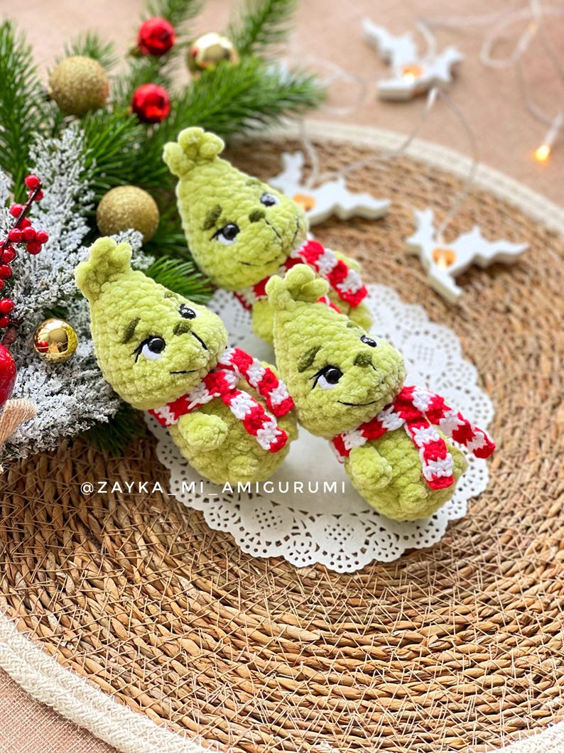 May include: Three handmade green crochet figures with black eyes and red and white striped scarves. The figures are arranged on a white lace doily, with Christmas ornaments and greenery in the background. The text "@ZAYKA_MI_AMIGURUMI" is visible.